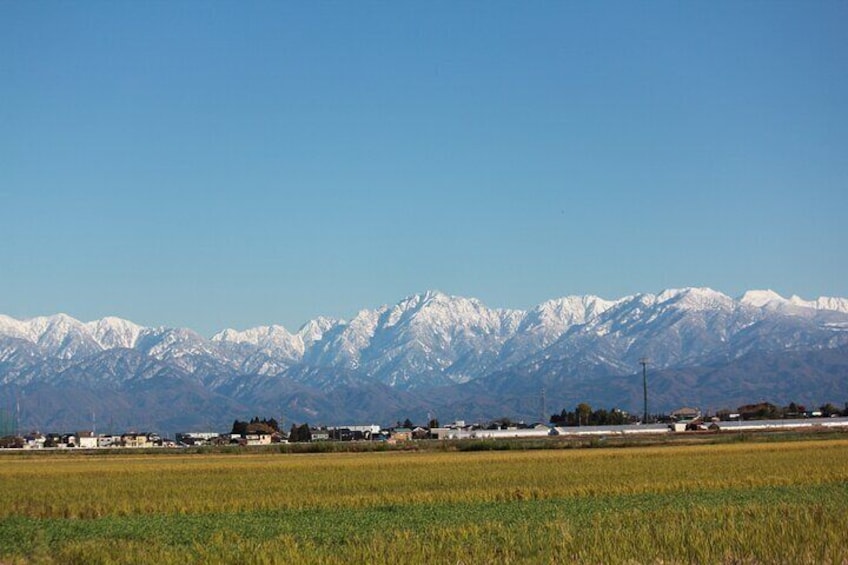 A mountain range with snow-capped peaks, a flat agricultural field, and a few buildings in the foreground. 1
