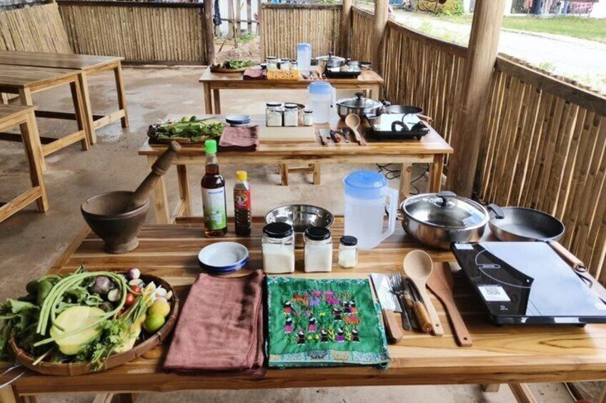 An outdoor dining setup with wooden tables, various food items, cooking utensils, and a portable stove. 5