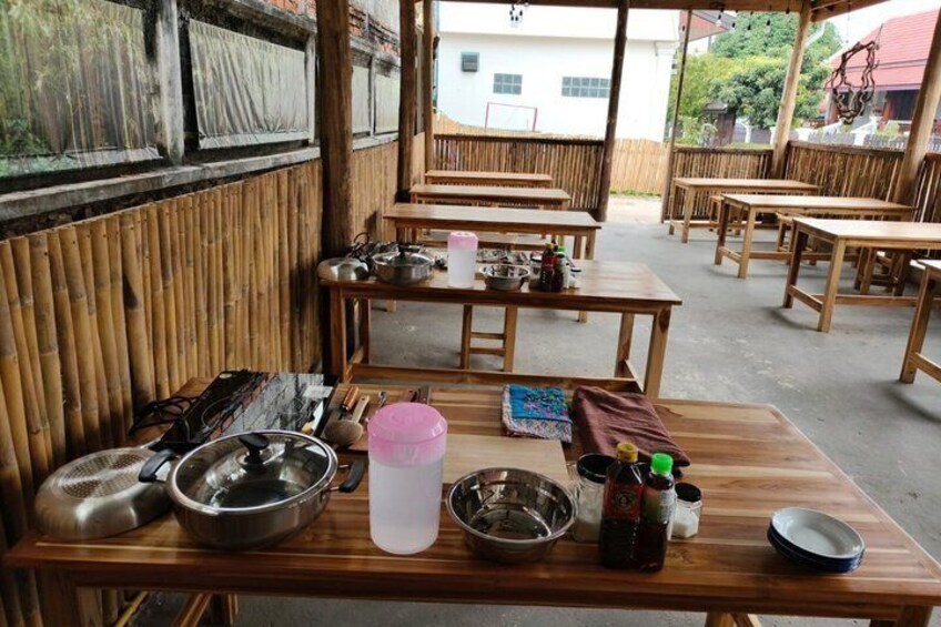 A bamboo-walled outdoor dining area with wooden tables and chairs, kitchen utensils, and a variety of dishes. 2