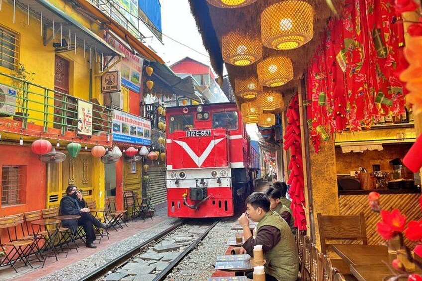 A red train on tracks with people dining outside a restaurant. 4