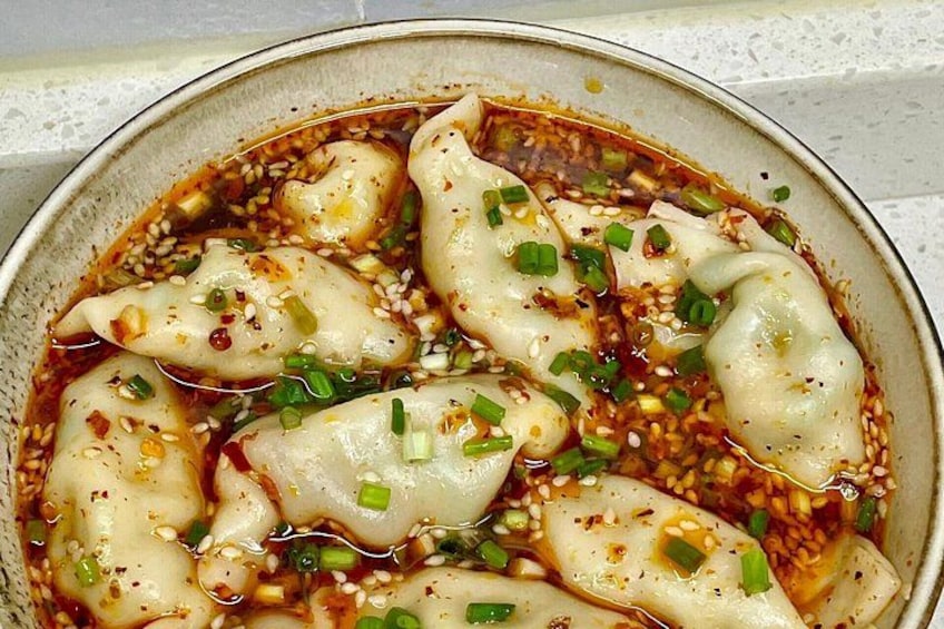 A bowl of dumplings in a sauce with sesame seeds and chopped green onions. 2