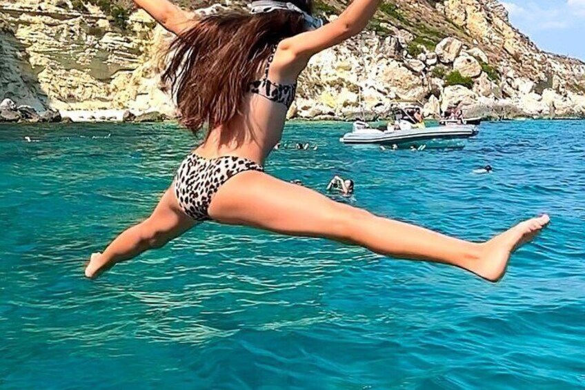A person jumping into turquoise water with a rocky coastline and a boat in the background. 5