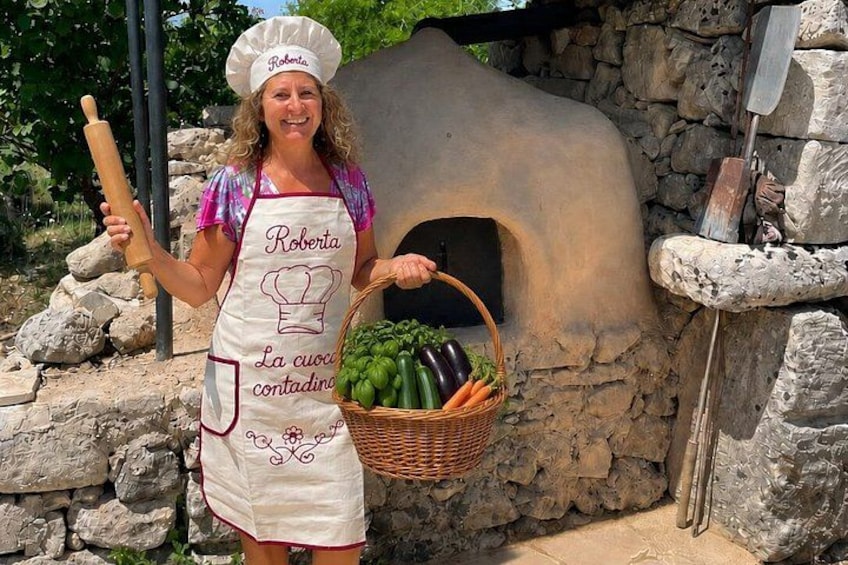 A person with a chef's hat and apron holding a rolling pin and a basket of vegetables in front of a stone oven. 1
