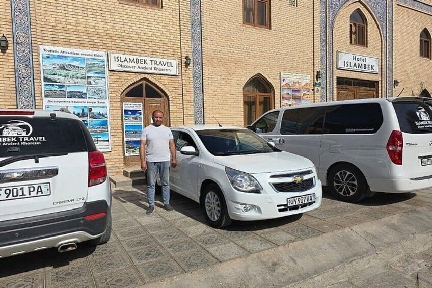 A man standing in front of a building with signs for 'Islambek Travel' and 'Hotel Islambek.' There are two white cars parked in front. 5