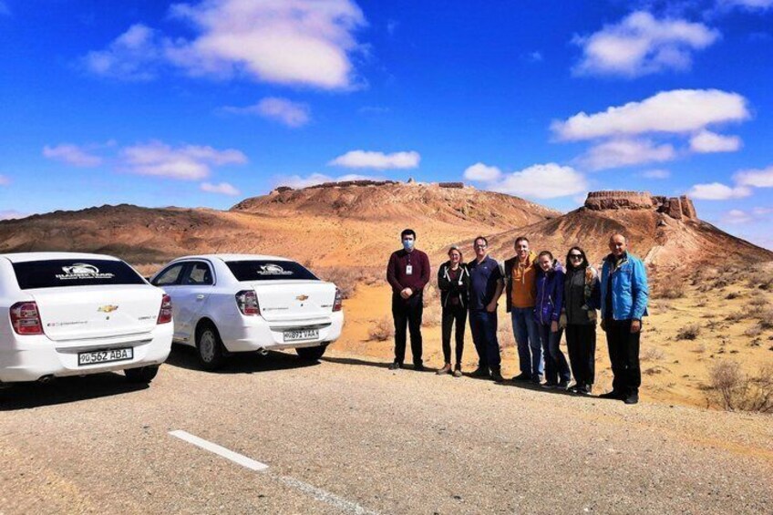 A group of people standing on a road with two white cars parked on the side, a desert landscape with hills in the background. 1