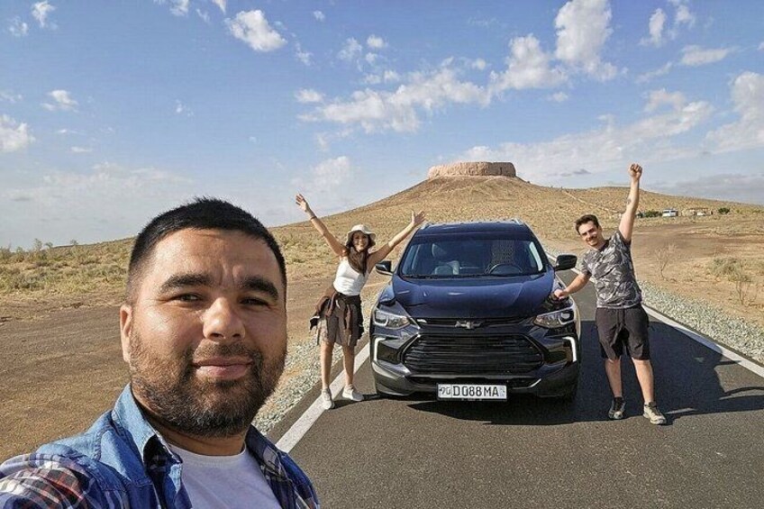 A desert landscape with a large mound in the background and a blue car parked on the roadside. 1