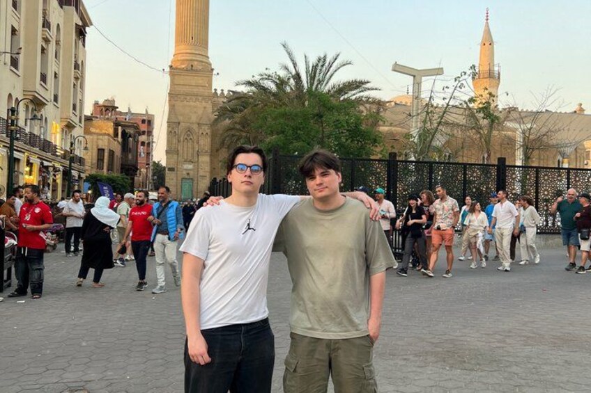 Two people standing in a public square with historical buildings and a minaret in the background. 5
