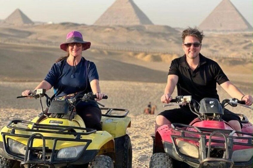 Two people on ATVs in a desert with pyramids in the background. 1
