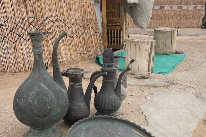 A collection of antique vases and a teapot on a dirt surface with a bamboo fence and wooden stools in the background. 2