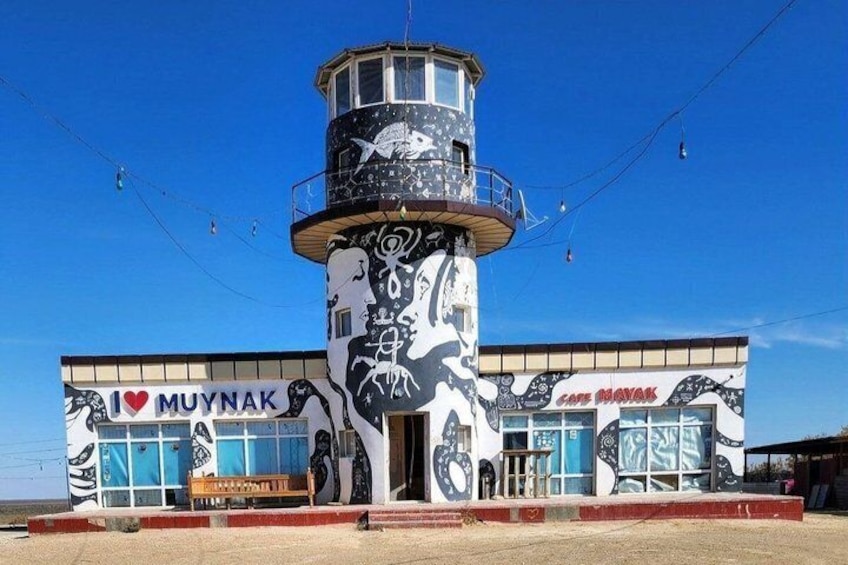 A building with a lighthouse tower and murals, labeled 'I ♥ MUYNAK' and 'Café MUYNAK.' 1