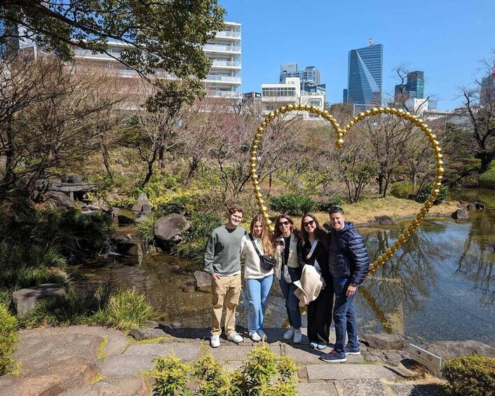 A group of people posing with a heart-shaped golden decoration in a park with a pond and buildings in the background. 3