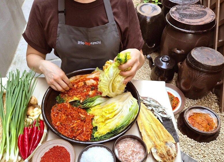 A person in a brown apron holding a cabbage, with various Korean dishes and ingredients like kimchi, chili flakes, and seafood on the table. 1