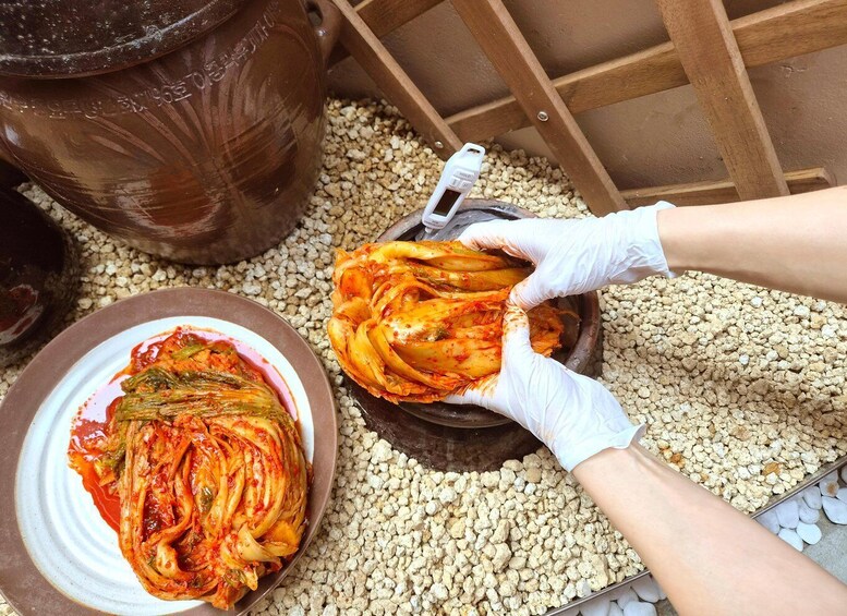 A person handling kimchi with gloved hands, surrounded by a large clay pot and a plate of kimchi. 4
