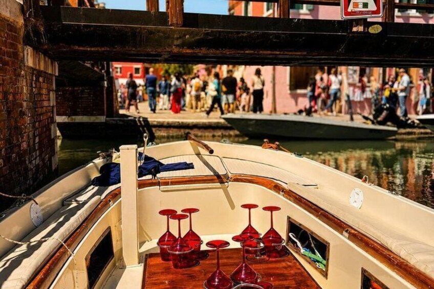 A narrow canal with a boat docked, red wine glasses on the deck, and people walking on the adjacent bridge. 4