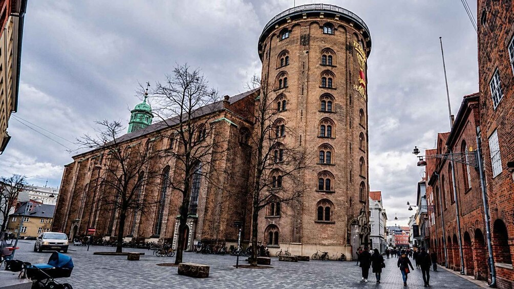 Ein historisches Gebäude mit einem runden Turm, einer grünen Kuppel und einem Pflasterplatz mit Fußgängern. 2