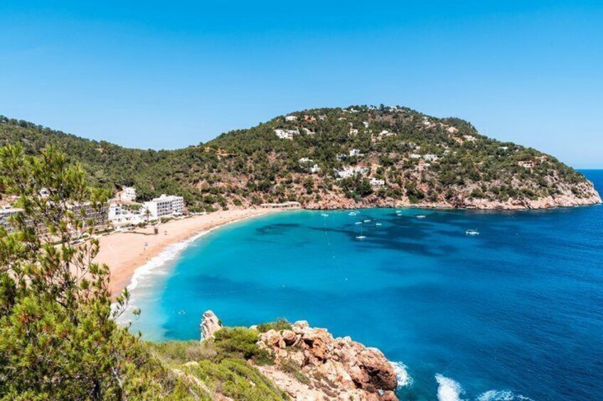 A coastal landscape with a beach, clear blue waters, and a hill with buildings. 1