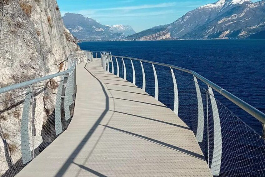 A coastal walkway with a metal railing, a paved path, and a view of the sea and mountains in the background. 2