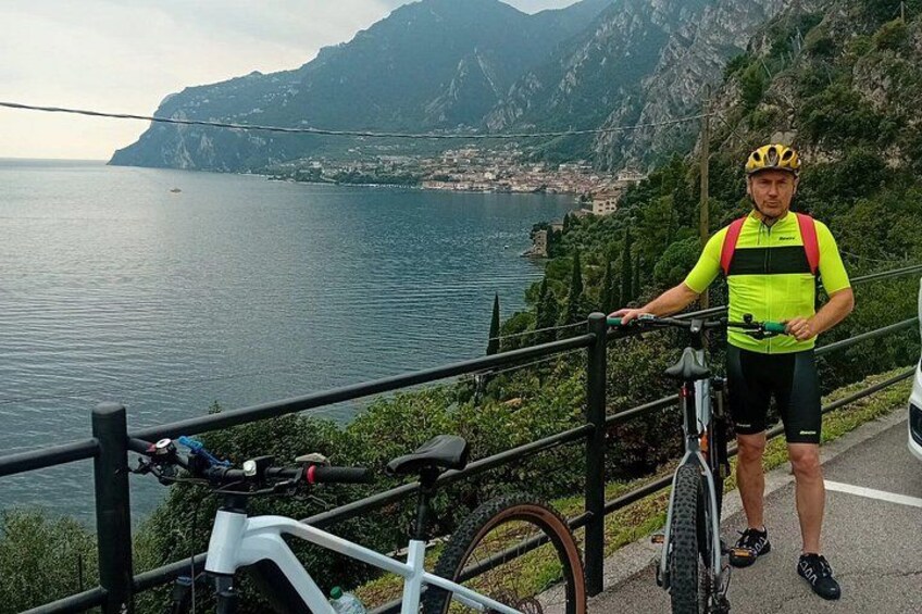 A cyclist standing next to a bicycle on a coastal road with a mountain backdrop. 1