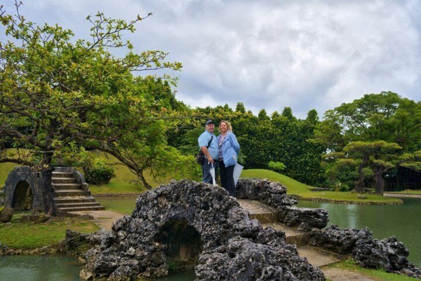 A couple standing by a pond with a stone bridge and lush greenery. 5