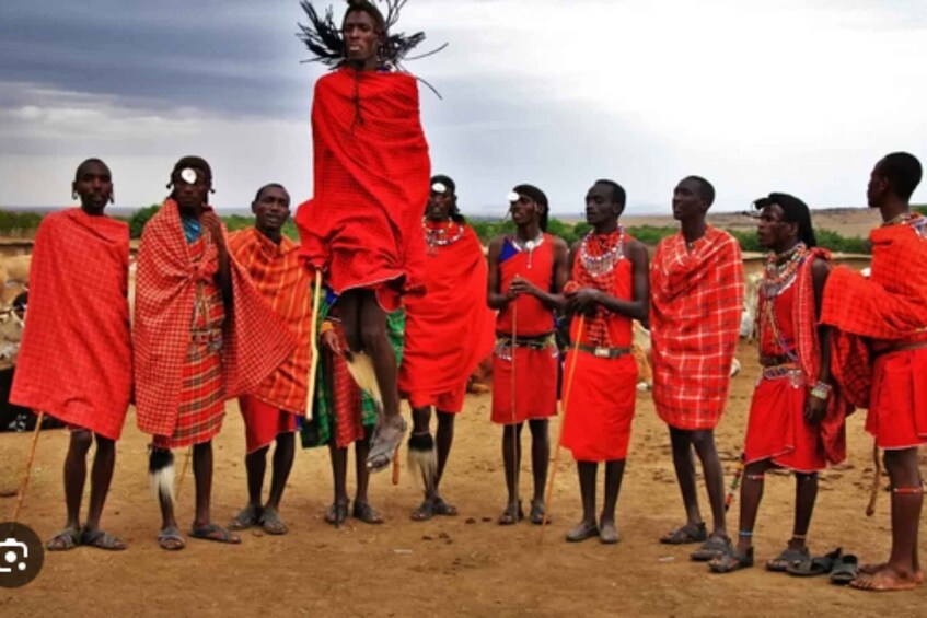 A group of people in traditional attire standing in a line on a dirt ground with a cloudy sky in the background. 2