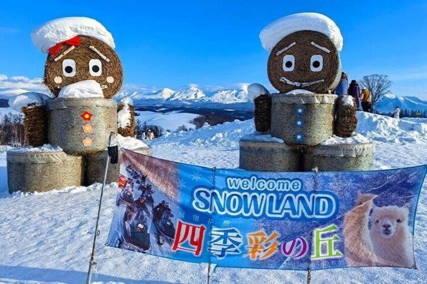 A snow-covered landscape with two large sculptures resembling people wearing winter hats and holding a barrel. A banner in the foreground reads 'Welcome to Snowland.' 2