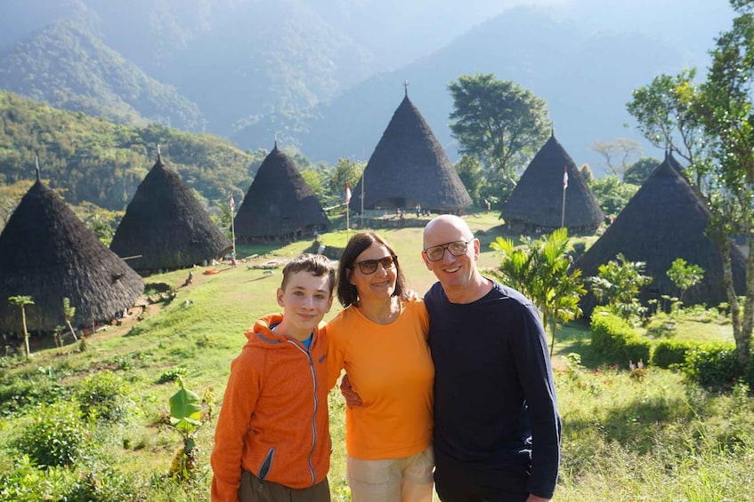A family standing in front of traditional conical huts with a mountainous backdrop. 1