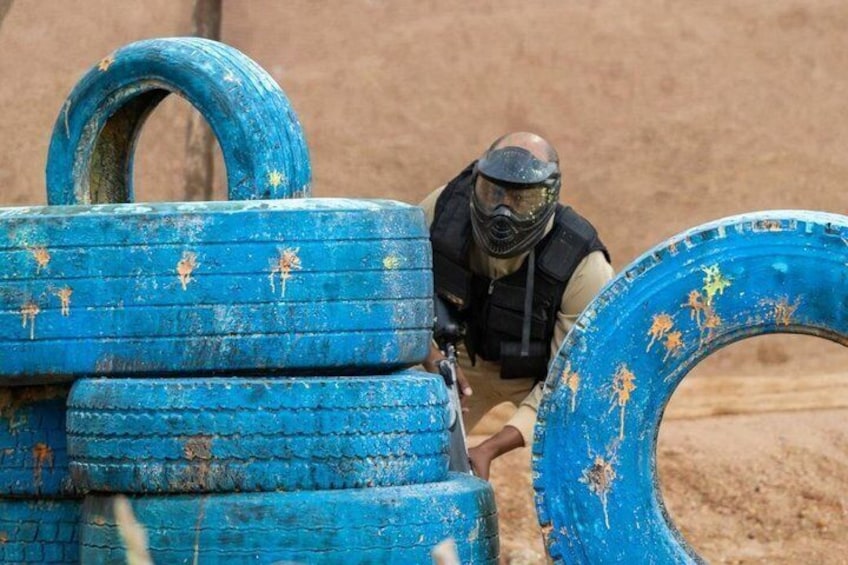 A person in tactical gear and a helmet, crouching behind blue tires in a desert-like environment. 3