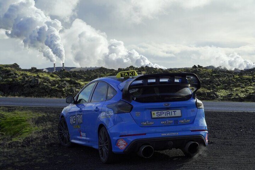 A blue rally car with a 'TAXI' sign on the roof, parked on a road with a backdrop of industrial smokestacks and a cloudy sky. 5