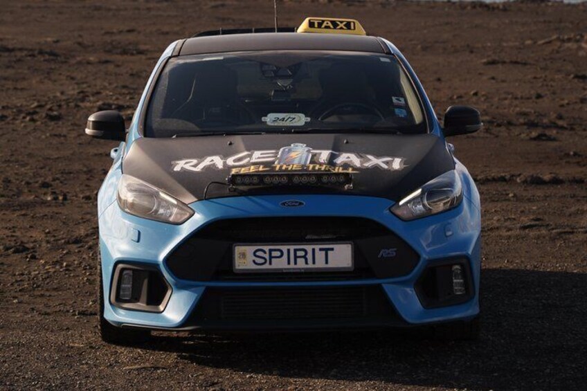 A blue car with 'RACE TAXI' and 'SPIRIT' on the front, parked on a dirt surface. 1