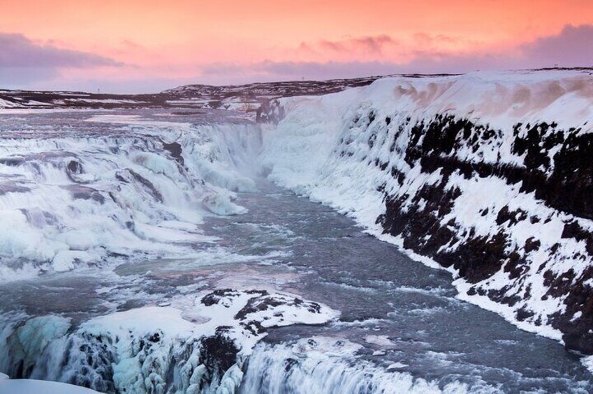 A waterfall cascading over a rocky cliff with snow patches, under a pink sky. 5