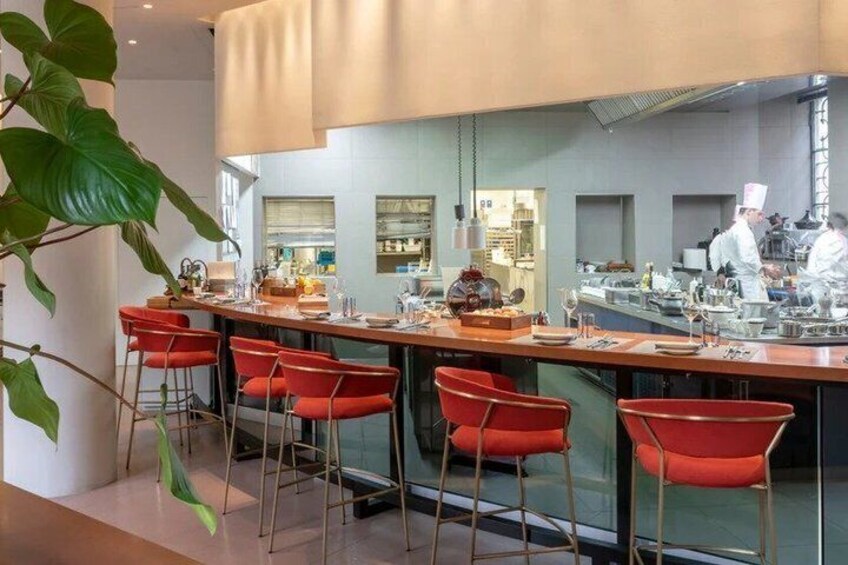 A modern restaurant with red bar stools, a kitchen in the background, and a large plant. 2
