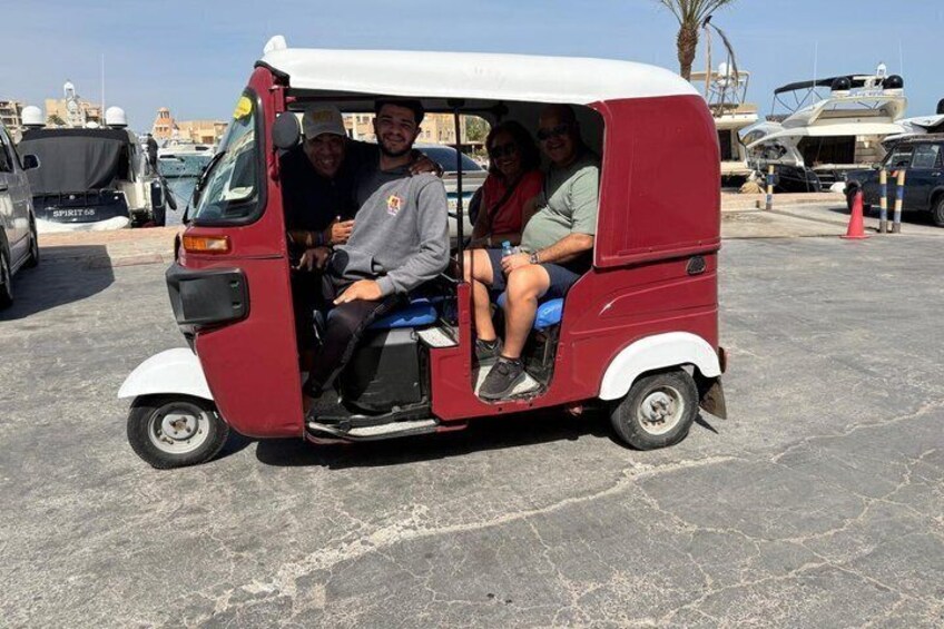 A red and white tuk-tuk with three passengers, parked on a paved surface with boats and yachts in the background. 4