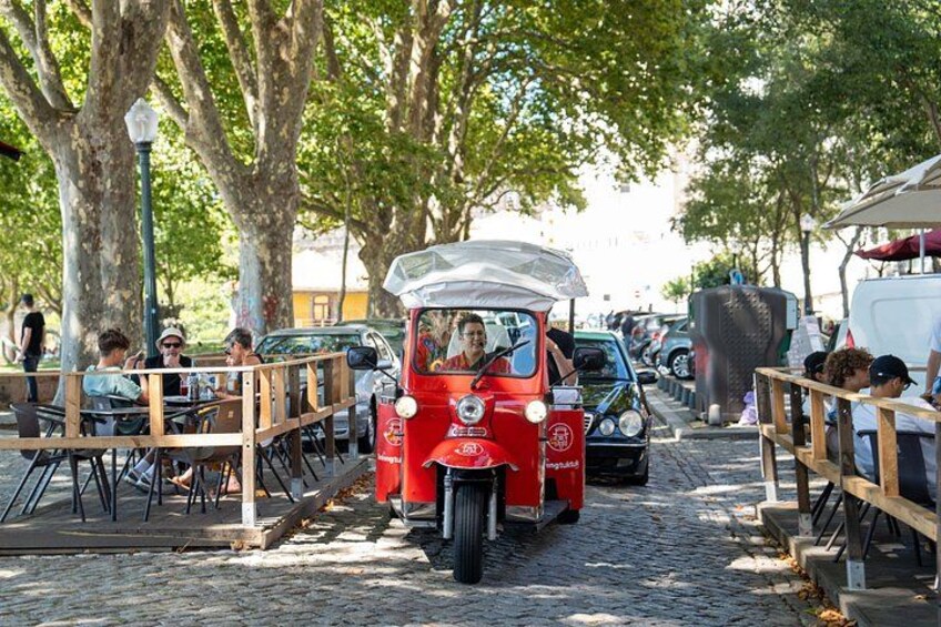 A red vintage car driving down a cobblestone street lined with trees and outdoor seating areas. 3
