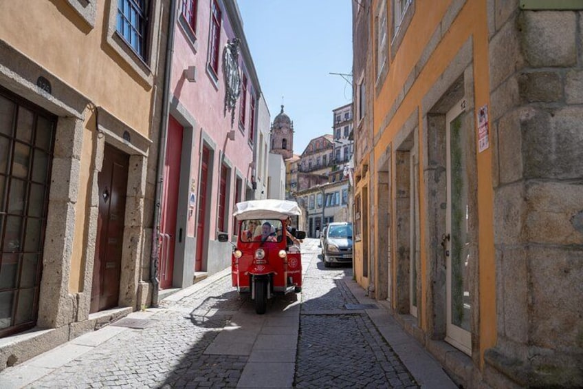 A narrow cobblestone street with historic buildings, a red vintage car, and a blue car parked on the side. 1
