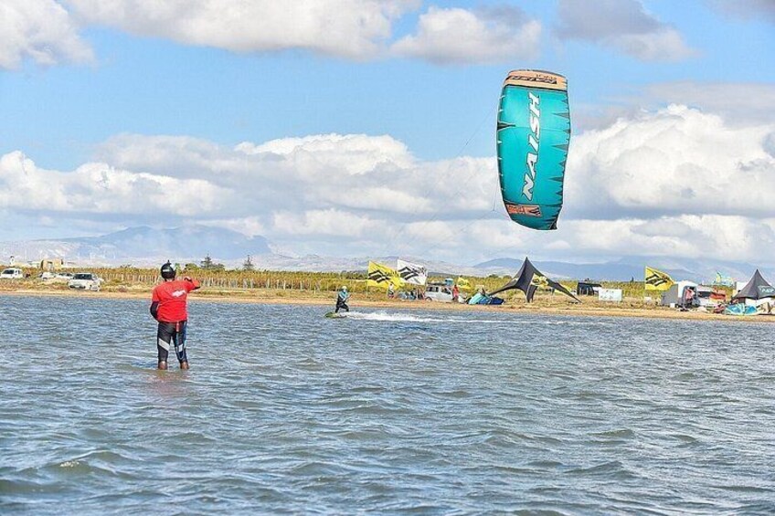 A person kiteboarding on water with a blue kite, another person on a board, and tents in the background. 3