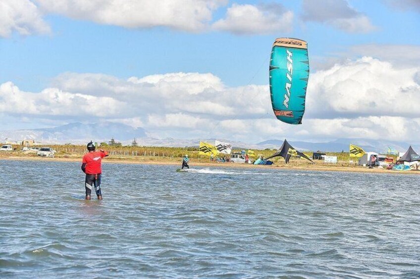 A person kiteboarding on water with a blue kite, another on a board, and tents in the background. 3