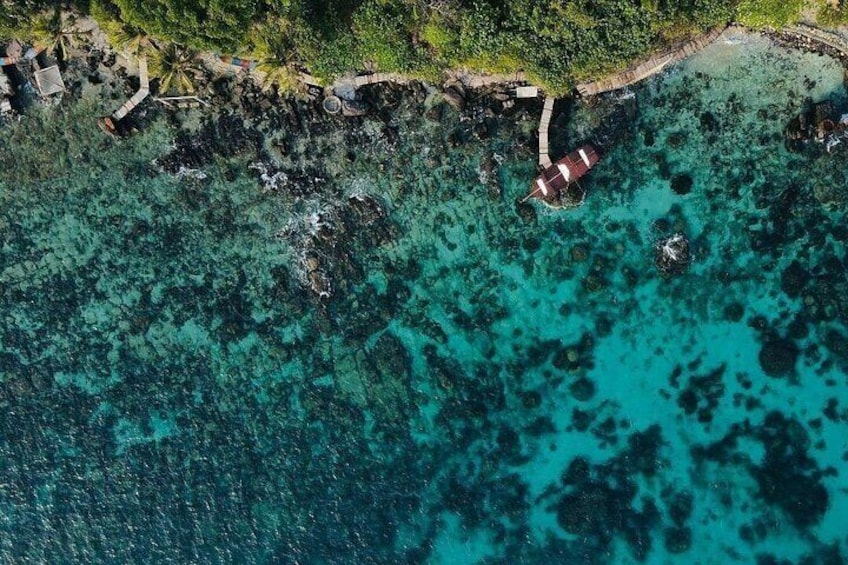 An aerial view of a coastal area with clear turquoise waters, rocky outcrops, and a small structure with a red roof near the shore. 3