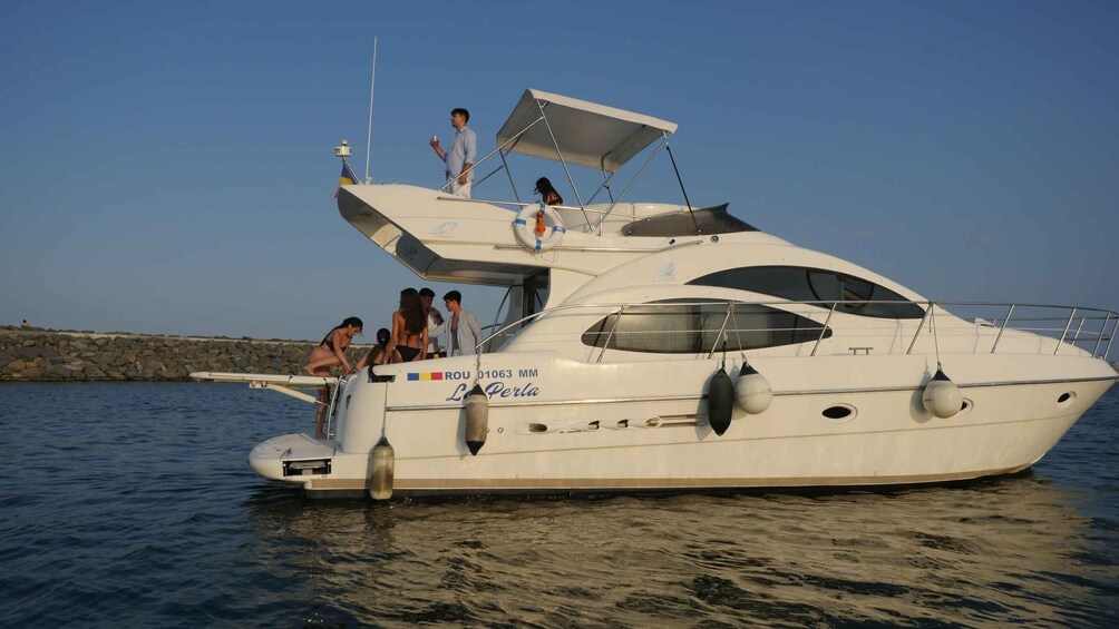 A white yacht named 'L'Erve Perla' with a partially open canopy, anchored near a rocky shoreline. 1