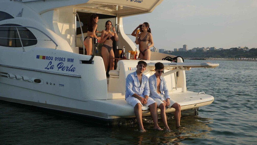 A yacht named 'La Perla' with three people on board, two seated and one standing, near a body of water with a cityscape in the background. 4