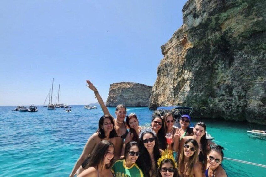 A group of people on a boat near a rocky coastline with clear blue water and sailboats in the distance. 4