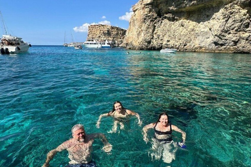 Three people swimming in clear blue water with boats and rocky cliffs in the background. 3