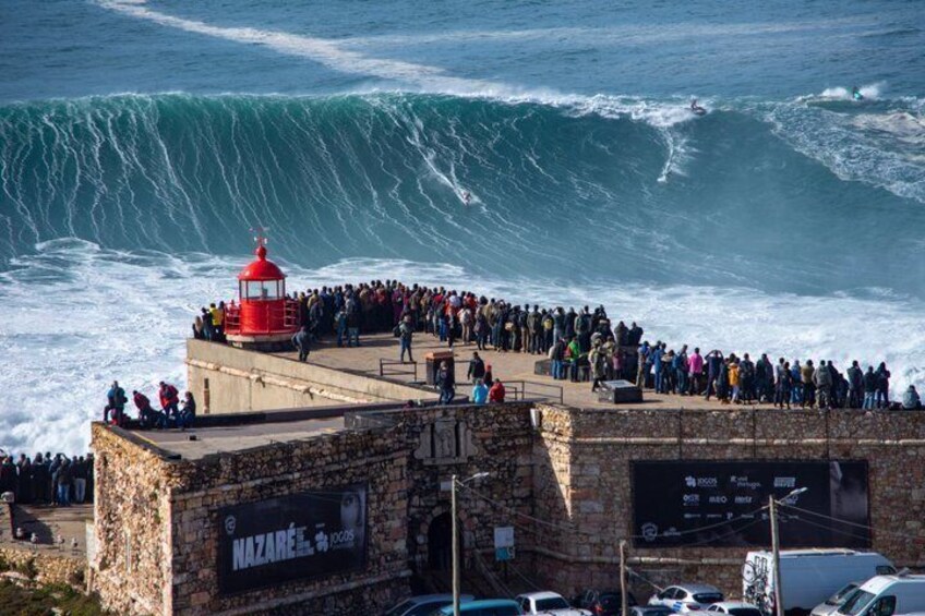 A coastal scene with a large crowd watching a massive wave, a red lighthouse, and a stone building with signage. 1