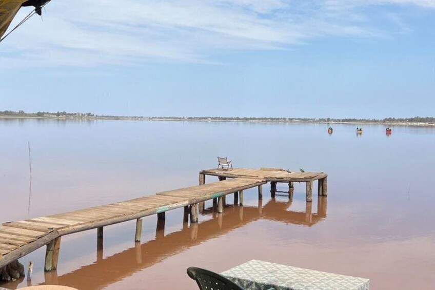 A wooden pier extending into calm water with a chair and a table on it, and a rowboat in the distance. 3