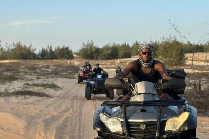 A person riding an ATV on a dirt road with sparse vegetation in the background. 1