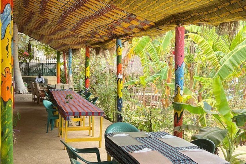 A covered outdoor seating area with colorful painted columns and tables, surrounded by lush banana plants. 5