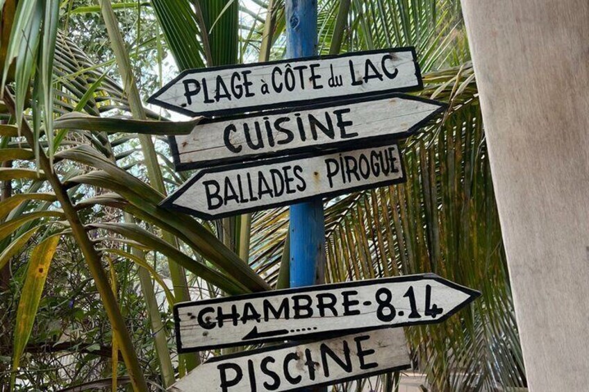 A directional signpost with multiple arrows pointing to various destinations such as 'Plage à Cote du Lac,' 'Cuisine,' 'Ballades,' 'Pirogue,' 'Chambre 8.14,' and 'Piscine.' 4