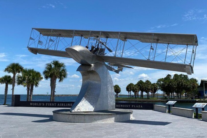 A large sculpture of a biplane with a circular base, located in an open plaza with palm trees and a view of the ocean. 4