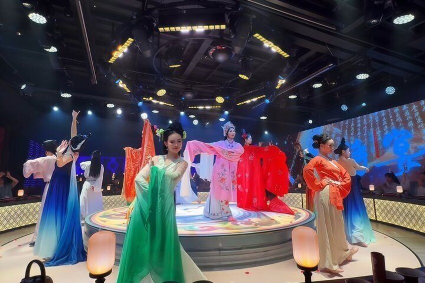 A stage with performers in traditional costumes, a circular platform, and a large screen in the background. 3
