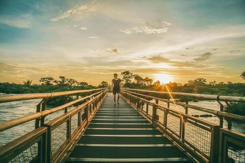 A wooden bridge with metal railings leading to a sunset over water. 2