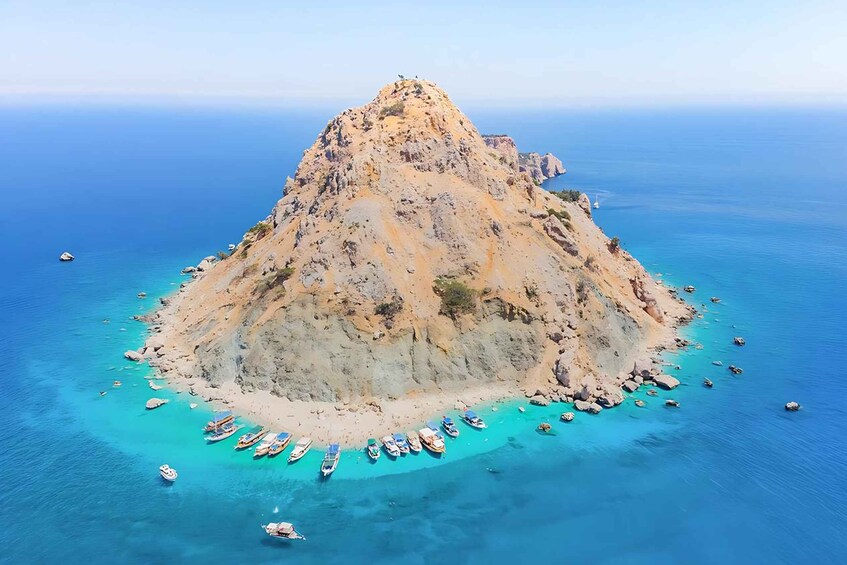 A rocky island surrounded by clear turquoise waters with several boats anchored near the shore. 3